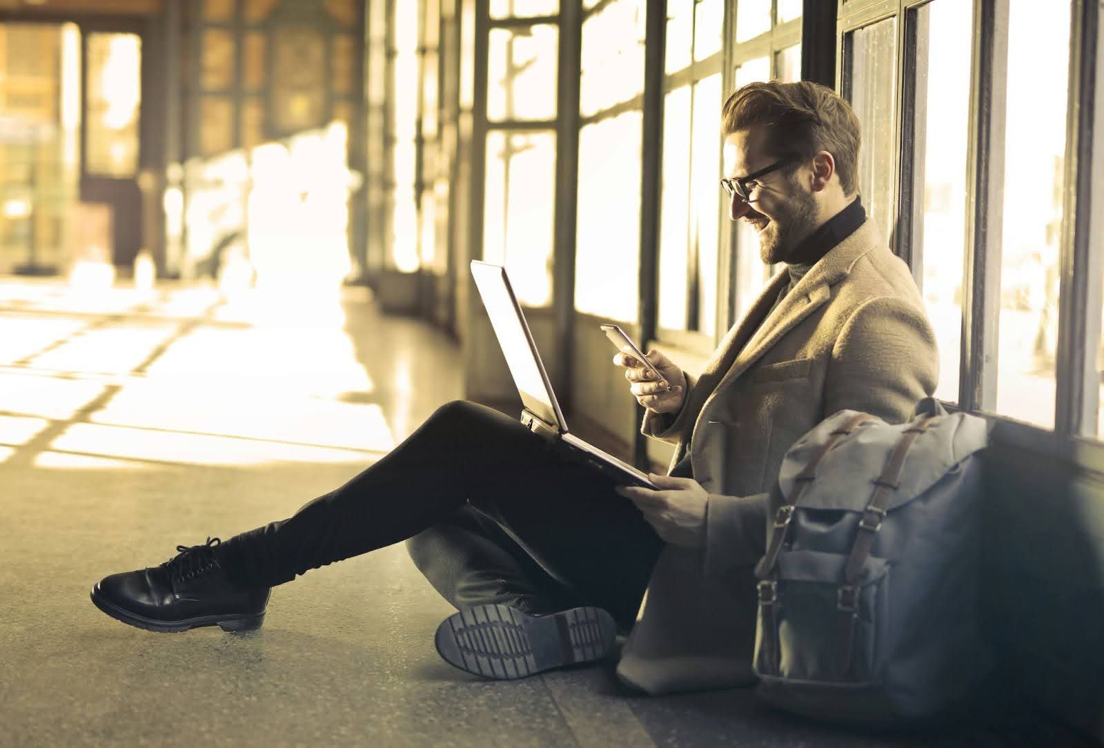 Man sitting by a window using edtech tool on a laptop and phone, with a backpack beside him, in a sunlit room.