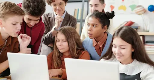 Group of students collaborating on laptops during a classroom jeopardy with teacher overseeing.